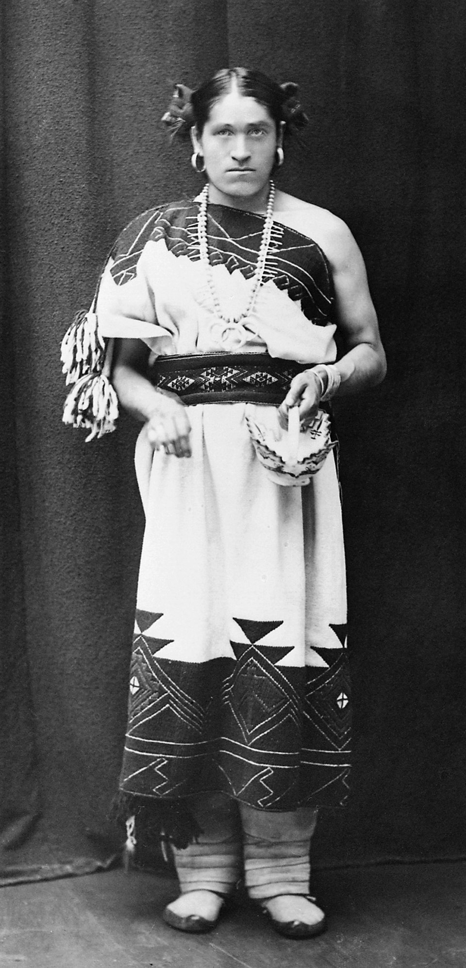 A black-and-white portrait of We’wha, a 19th-century Zuni lhamana, standing against a dark backdrop. She wears a traditional woven Zuni dress decorated with geometric patterns, a belted sash at the waist, and layered necklaces of shell and stone. Her hair is parted and styled in rounded side knots, a form associated with Zuni women’s hairstyles of the period. She holds a small woven basket in one hand and stands with wrapped moccasins on her feet. The image reflects her role as a skilled potter, weaver, and